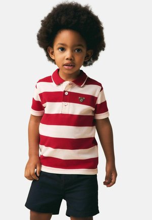 Noor laps lokkis juustega, seljas punase ja valge triibulise poloshirt karuembleemiga ning tumesinised lühikesed püksid, seisab ühevärvilise tausta ees.