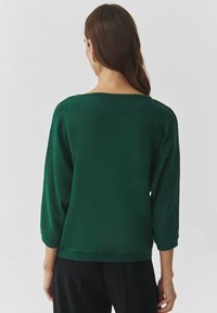 Maglione verde lavorato a maglia con maniche a tre quarti, caratterizzato da piccole perline lungo le cuciture delle maniche e un ampio scollo. Texture morbida.