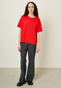 Raudona trumpomis rankovėmis marškinėliai, laisvas siluetas, derinami su pilkomis plačiomis kelnėmis ir juodais masyviais batais, prieš neutralią foną.