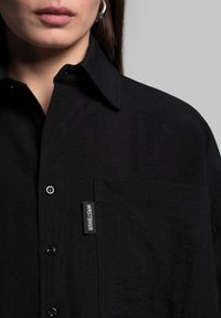 Zwarte overhem shirt van textuurstof. Beschikt over een zak met een logo-label en een klassieke kraag. De knopen zijn donker en rond.
