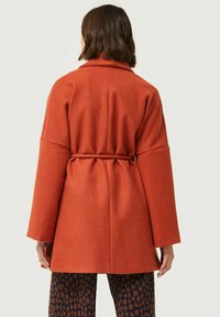 Abrigo envolvente de lana naranja con mangas anchas y un cinturón en la cintura, de tela texturada, visto desde atrás. Combinado con pantalones con estampados.
