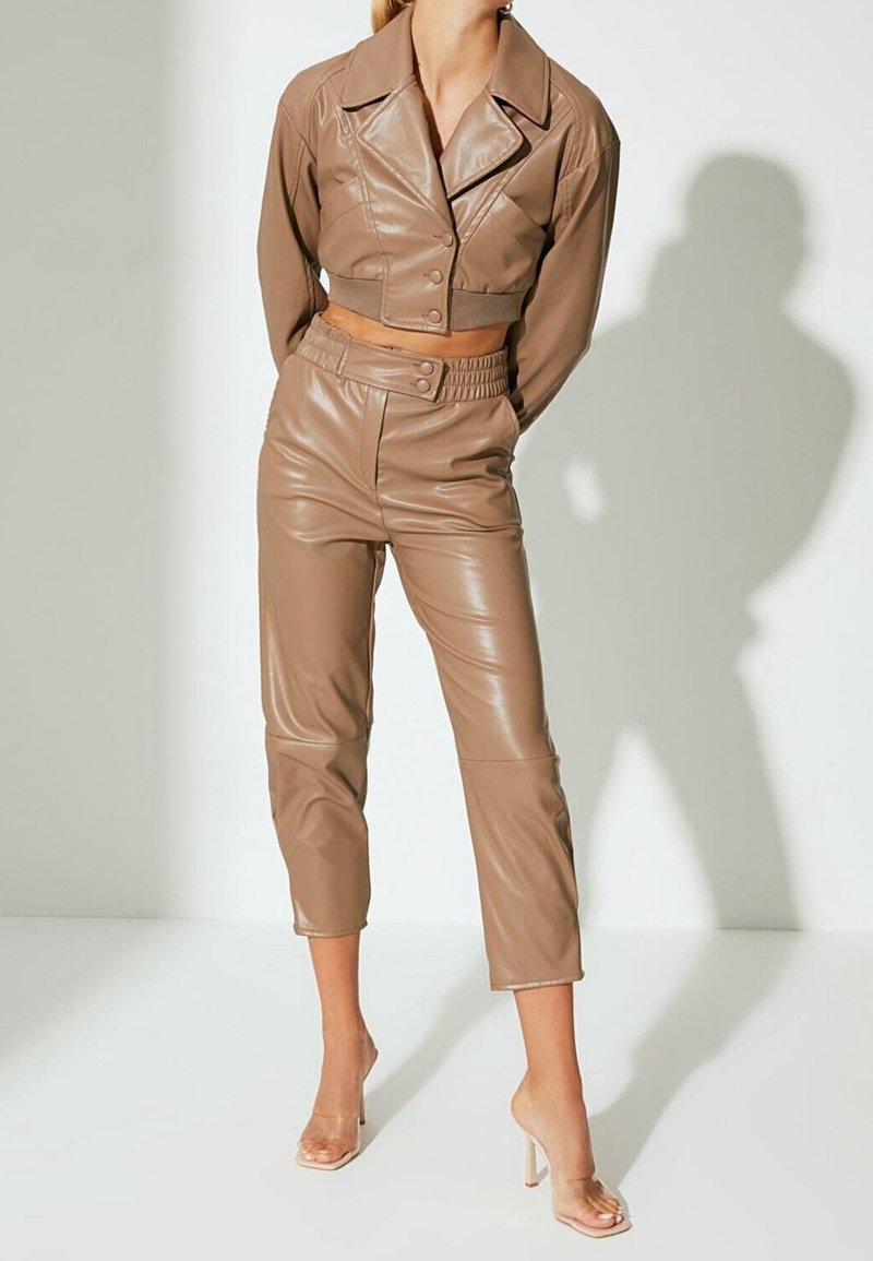 Braune Kunstleder-Cropped-Jacke mit einem Kragen und Knopfverschluss, kombiniert mit hoch taillierten, passenden schmalen Hosen. Durchsichtige, hochhackige Sandalen.
