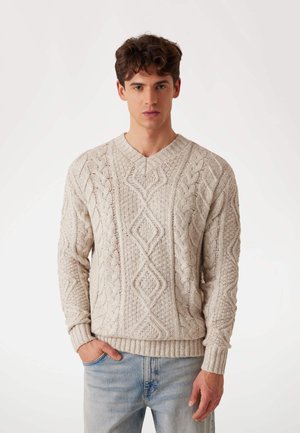 Light beige knitted sweater with a textured cable pattern, V-neck design, and ribbed cuffs and hem, paired with light blue jeans.