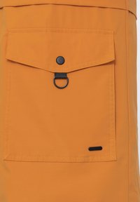 Poche en tissu orange avec bouton-pression noir, boucle en D noire et petite étiquette rectangulaire noire dans le coin inférieur droit.