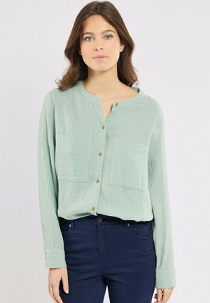 Jeune femme aux cheveux bruns portant un chemisier vert clair à boutons et un pantalon bleu foncé, debout devant un fond uni.