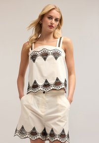 Top alb fără mâneci cu un model brodat în dungi negre și accente maronii, asociat cu pantaloni scurți albi care au margini cu un model similar.