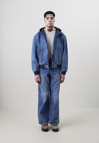 Denim jakna s kapuljačom, svijetloplave boje i prednjim dzepovima; uparena s širokim trapericama u istom tonu; bež cipele upotpunjuju odjevnu kombinaciju.