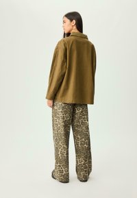 Žena s dugom tamnom kosom nosi preveliki maslinasti kaput, široke hlače s leopard uzorkom, naušnice na obruč i crne cipele, stoji okrenuta leđima.