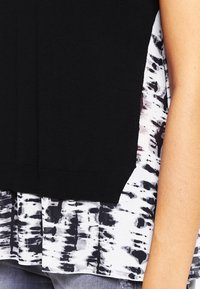 Musta yläosa, jossa on teksturoitu neulottu yläosa ja valkoinen, laskeutuva kangas alaosassa, jossa on musta abstrakti kuvio. Sauma näkyy vyötäröllä.