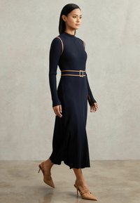 Marineblauwe ribgebreide jurk met lange mouwen en een taille met ceintuur, met een aansluitend lijf, een uitlopende rok en zijsplitten. Gecombineerd met beige puntige hakken.