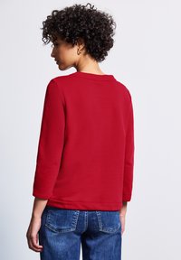 Röd långärmad topp med rund halsringning och avslappnad passform, kombinerad med blå denimjeans med bakfickor och en lätt utsvängning.