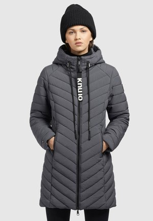 Femme portant un manteau d'hiver matelassé gris avec capuche et fermeture éclair à l'avant, les mains dans les poches, contre un fond uni.
