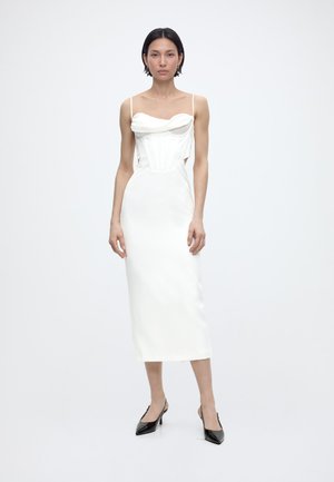 Kvinde med kort mørkt hår iført en hvid ærmeløs midikjole med udskæringer, parret med sorte spidse slingback-hæle, stående mod en ensfarvet baggrund.