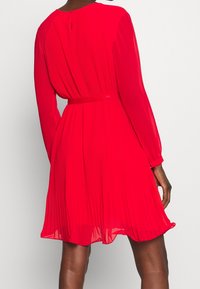 Nainen, jolla on päällään kirkkaan punainen, laskostettu polvipituinen mekko, jossa on pitkät hihat ja kapea vyö, nähtynä takaa yksinkertaista taustaa vasten.