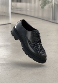 Chaussure Oxford en cuir noir et tweed avec une semelle en caoutchouc épaisse, flottant au-dessus d’un sol en béton poli dans un intérieur moderne.
