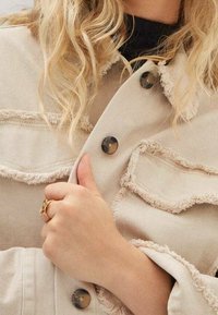 Beige denimjakke med slidte kanter, to brystlommer og runde knapper. En hånd med en ring holder jakken ved kraven.