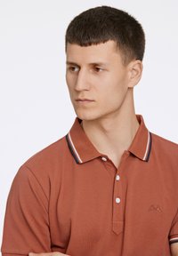 Polo de couleur rouille avec une finition texturée, doté d'une patte de boutonnage à trois boutons et d'un col rayé bleu marine et blanc. Logo brodé sur la poitrine.