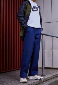 Giacca Nike nera e oliva, con logo bianco, abbinata a un felpa bianca. Pantaloni navy larghi e sneakers bianche con dettagli multicolori.