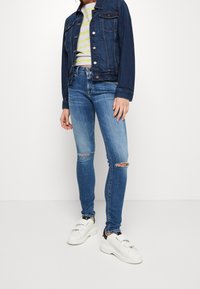 Jeansowa kurtka, ciemnoniebieska, zapinana na guziki, z dwiema kieszeniami na piersi. Szary prążkowany crop top. Niebieskie skinny jeans z dziurami na kolanach. Białe sneakersy z czarnymi akcentami.