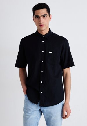 Black short-sleeve button-up shirt with a chest pocket, featuring white buttons and a small brand label on the pocket. Smooth texture.