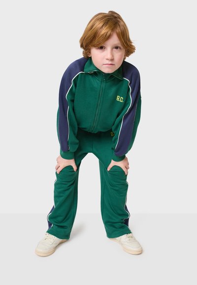 Zelený a námornícky modrý teplákový súbor s bielymi akcentmi, ktorý obsahuje bundu na zips a nohavice s elastickým pásom, vyrobený z mäkkej, textúrovanej látky.