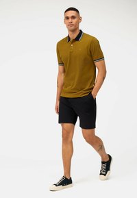 Gelbes Poloshirt mit marineblauem Kragen und Streifenakzenten, kombiniert mit schwarzen Shorts und dunklen Sneakern. Das Model steht selbstbewusst mit den Händen in den Taschen.