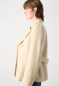 Béžový dvojradový kabát z vlnenej zmesi so zárezovým golierom, bočnými vreckami a detailom v páse s opaskom. Hladká textúra, štruktúrovaný strih.