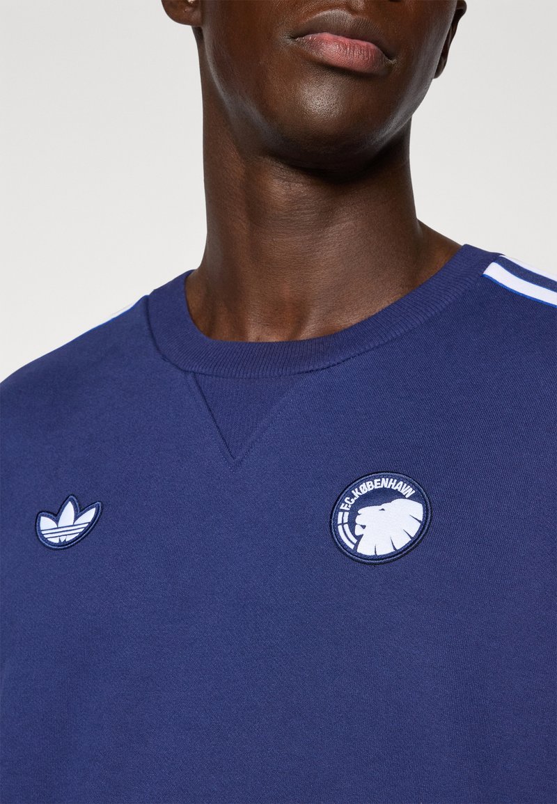 Sudadera azul marino con cuello acanalado; presenta un logotipo bordado de Adidas y una insignia del FC Copenhague en el pecho; tres franjas blancas en las mangas.