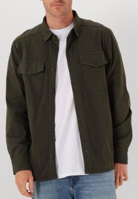 Man in een donkergroen overhemd met knopen over een wit T-shirt, met blauwe jeans, staand tegen een effen lichte achtergrond.