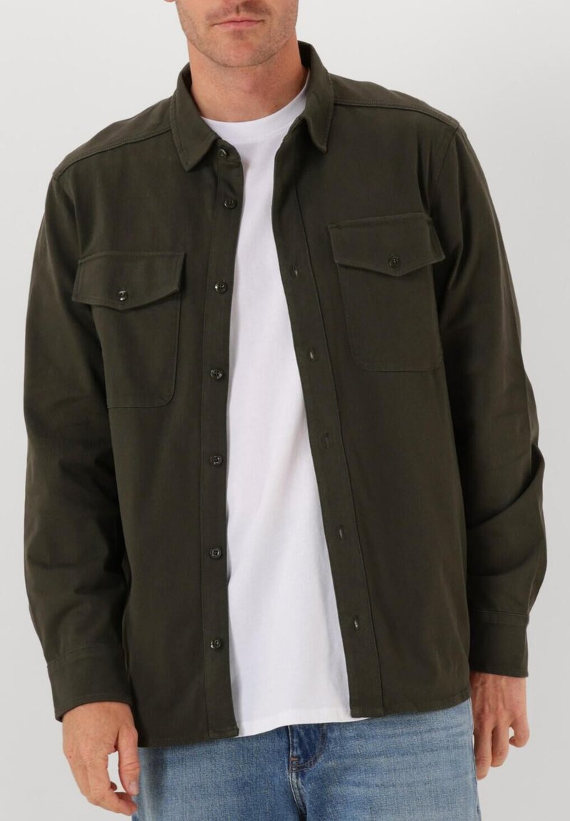 Man in een donkergroen overhemd met knopen over een wit T-shirt, met blauwe jeans, staand tegen een effen lichte achtergrond.