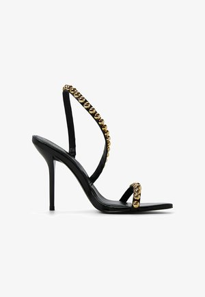 Sandalo nero con tacco alto e cinturino con perline dorate lucide. Presenta un design elegante e sottili cinturini alla caviglia e alle dita per supporto.