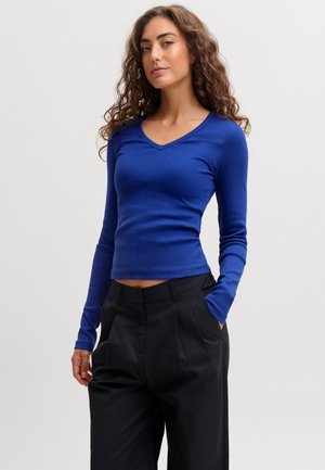 Femme aux cheveux bouclés portant un haut bleu ajusté à manches longues et un pantalon noir taille haute, debout avec une main dans la poche.