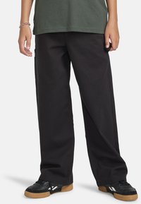 Zwarte wijde broek gemaakt van duurzaam katoen, met een relaxte pasvorm en naadloze zijzakken, gecombineerd met zwarte sneakers en een groene top.
