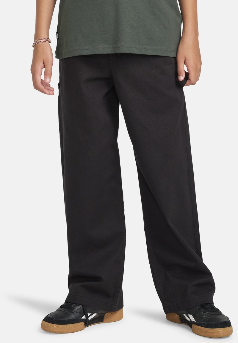 Zwarte wijde broek gemaakt van duurzaam katoen, met een relaxte pasvorm en naadloze zijzakken, gecombineerd met zwarte sneakers en een groene top.