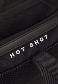 Czarny plecak tekstylny z wyściełanymi paskami i białym haftowanym napisem "HOT SHOT". Charakteryzuje się gładkim, trwałym materiałem i starannym szyciem.