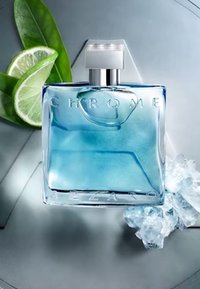 Bouteille de parfum en verre clair avec une forme rectangulaire. Liquide bleu clair, bouchon argenté et lettrage en relief "CHROME". Entourée de citron vert et de feuilles.