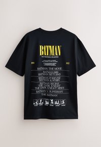 Camiseta negra de algodón con el texto amarillo llamativo "BATMAN" y una lista de películas en blanco, con detalles gráficos en la parte inferior y mangas cortas.