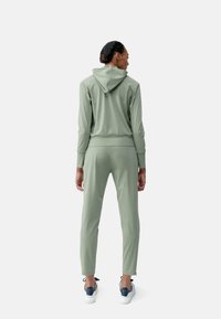 Completo di due pezzi di tuta leggera verde chiaro realizzato in tessuto morbido. Il cappuccio presenta una tasca frontale, mentre i pantaloni hanno dettagli con zip sulle caviglie.