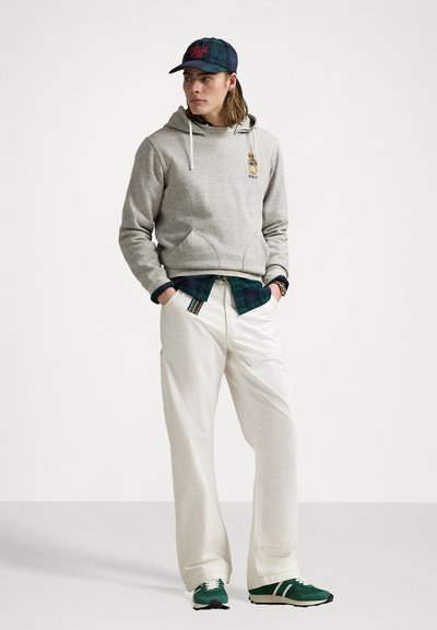Hoodie gris avec un personnage brodé, associé à une chemise à carreaux verte, un pantalon large blanc et des baskets vertes avec des accents blancs.
