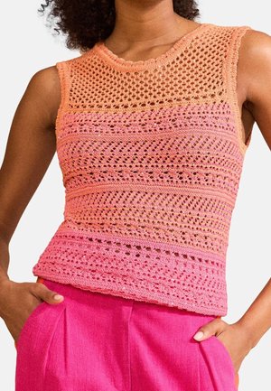 Bez-rękawnik z dzianiny na szydełku w gradientowych odcieniach pomarańczu i różu, noszony z różowymi, wysokowarstwowymi spodniami, ręce w kieszeniach.