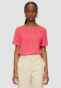 Korallefarbige Bluse mit kurzen Ärmeln und gefalteter Vorderseite, aus leichtem Stoff. Kombiniert mit beigen Hosen, glatte Textur, schlichtes Design.