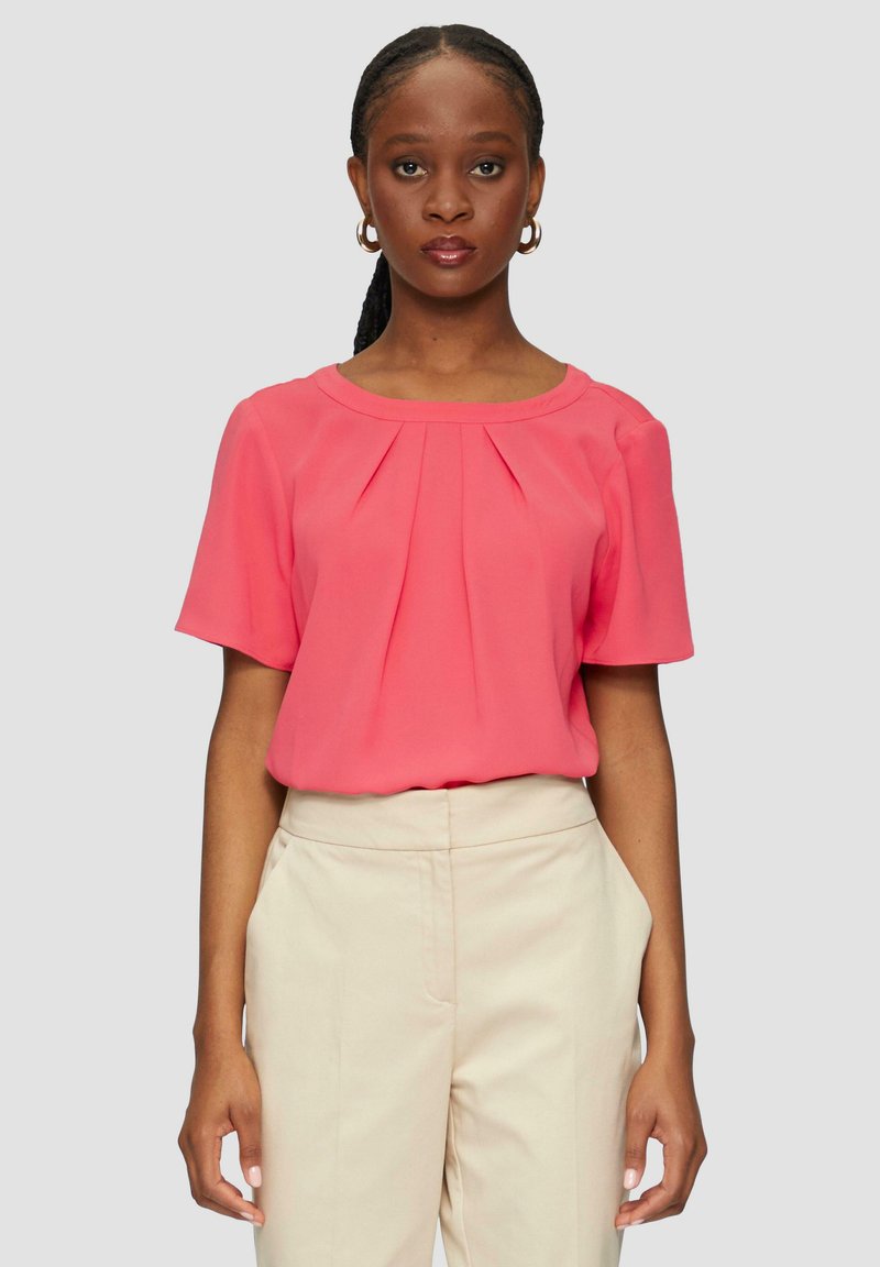 Korallefarbige Bluse mit kurzen Ärmeln und gefalteter Vorderseite, aus leichtem Stoff. Kombiniert mit beigen Hosen, glatte Textur, schlichtes Design.
