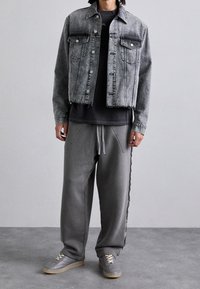 Džínová bunda světle šedé barvy se dvěma náprsními kapsami a roztrženým lemem, přetáhnutá přes černé tričko. Šedé kalhoty se šňůrkou a šedé tenisky.