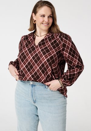Glimlachende vrouw met gouden oorringen, een bruin-roze geruite blouse en lichtblauwe spijkerbroek, poserend met handen in de zakken tegen een witte achtergrond.