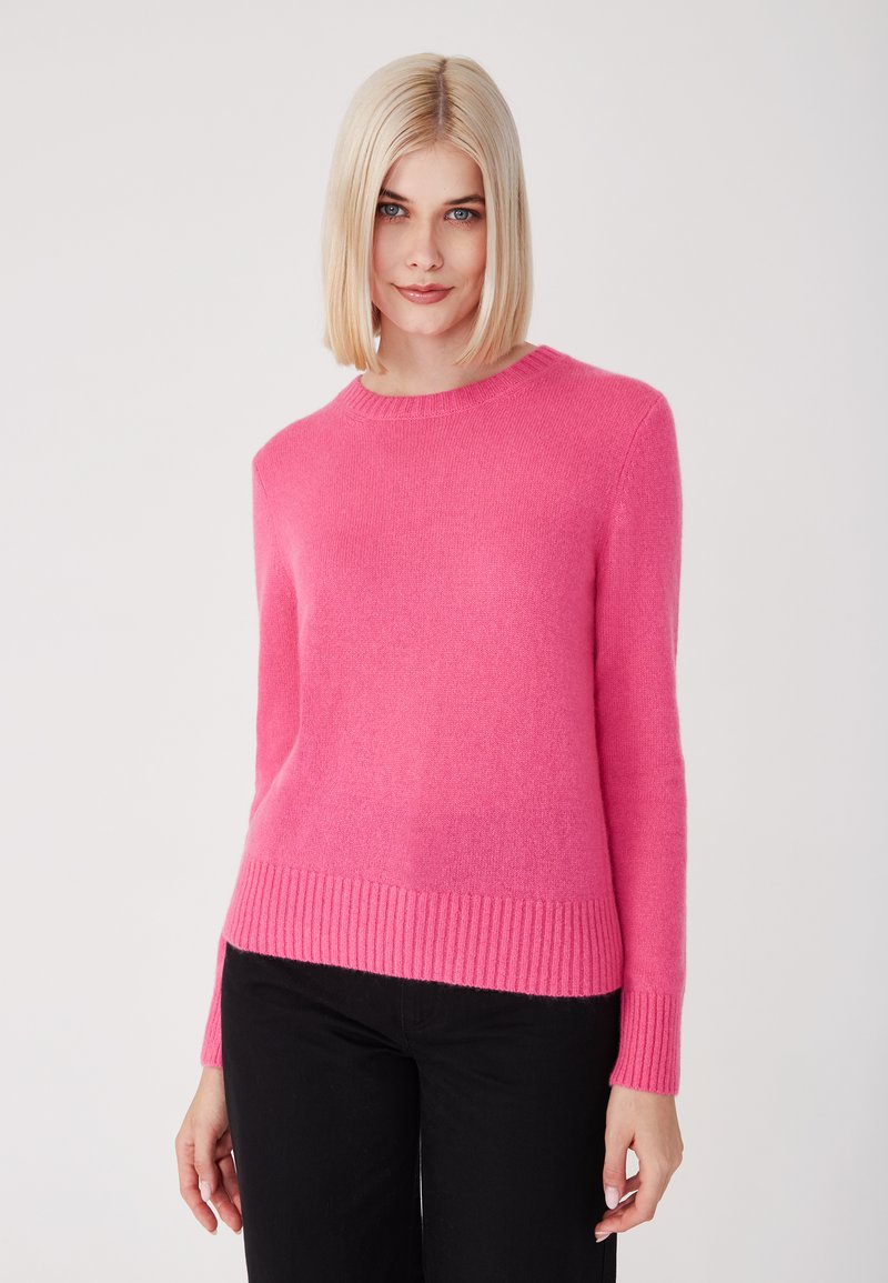 Suéter rosa de punto con cuello redondo, dobladillo y puños acanalados, que presenta una textura suave. Lleva puesto con pantalones negros.