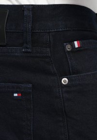 Tamsiai mėlyni džinsai iš denimo su lygia tekstūra, turintys mažą tricolorinį logotipą ir sidabro spalvos furnitūrą prie priekinio kišenės.