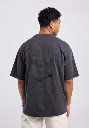 Jonge man met kort krullend haar, die een donker oversized T-shirt draagt met de tekst "Stay Wild Nature Club" en een vogelafbeelding op de achterkant.