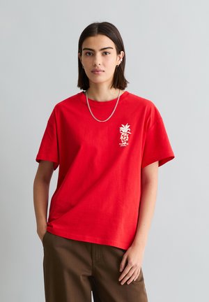 Kobieta z krótkimi ciemnymi włosami, ubrana w czerwoną koszulkę z nadrukiem, brązowe spodnie, łańcuszek na szyi i kolczyki, stojąca na tle jednolitego szarego tła.