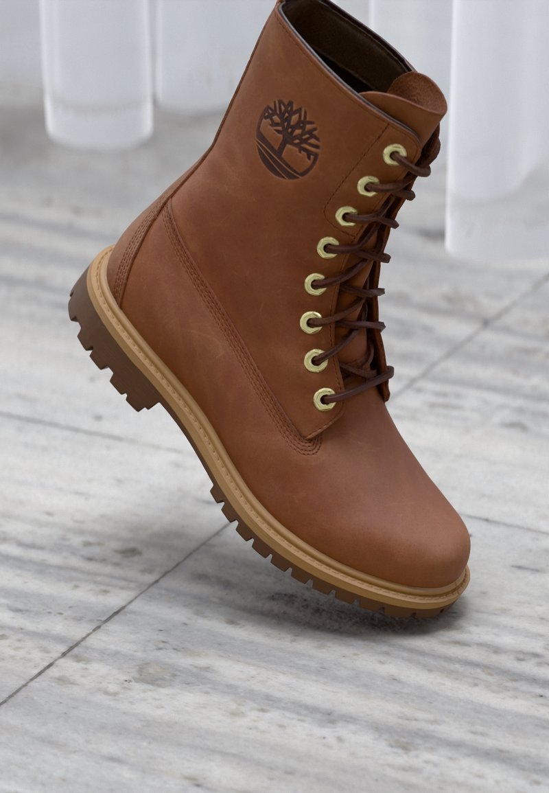 Botte en cuir marron à lacets avec semelle robuste et logo arbre, montrée inclinée sur un fond de sol en marbre clair.