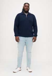 Σκούρο ναυτικό πουλόβερ πλεκτό με κολάρο μισού φερμουάρ, συνδυασμένο με ανοιχτό μπλε τζην και λευκά αθλητικά παπούτσια. Λείο υλικό και άνετη εφαρμογή.